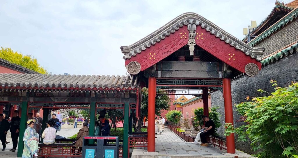 Kaiserpalast Shenyang – Die „Verbotene Stadt“ des Nordens erleben 19 Eine offene Arkade mit einem grauen Ziegeldach, roten Säulen und dekorativen Elementen, die zu einem Innenhof führt. Im Hintergrund sind weitere Palastgebäude und Menschen zu sehen.