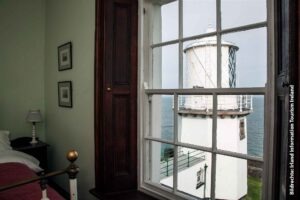 Blick aus einem gemütlich eingerichteten Zimmer mit hellgrünen Wänden durch ein weißes Sprossenfenster auf das strahlend weiße Blackhead Lighthouse und die Irische See unter einem bewölkten Himmel.