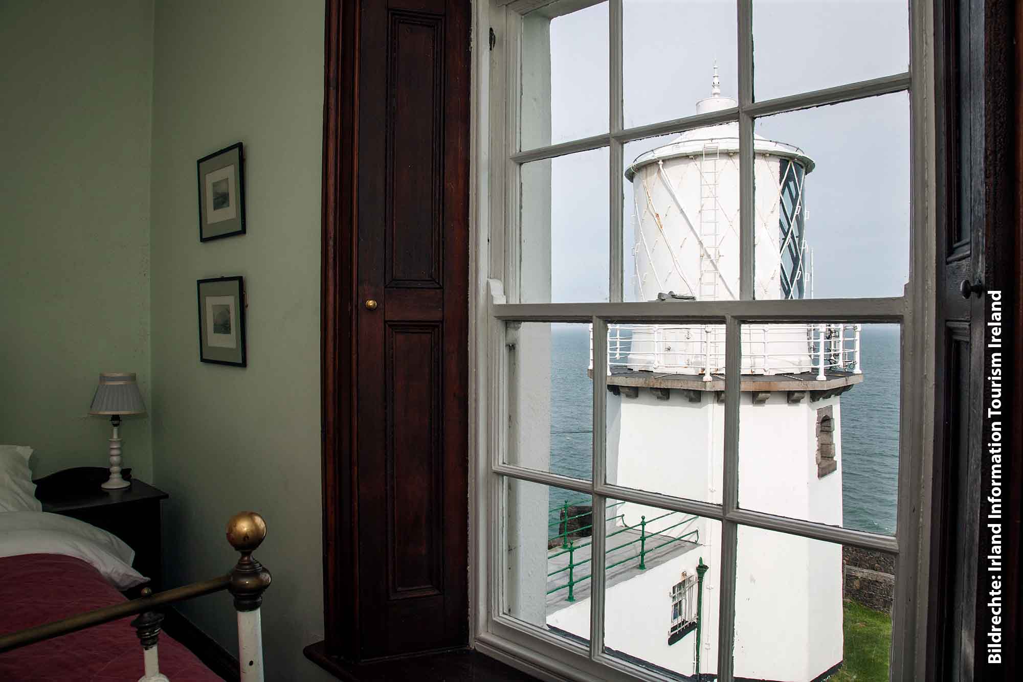 Blick aus einem gemütlich eingerichteten Zimmer mit hellgrünen Wänden durch ein weißes Sprossenfenster auf das strahlend weiße Blackhead Lighthouse und die Irische See unter einem bewölkten Himmel.