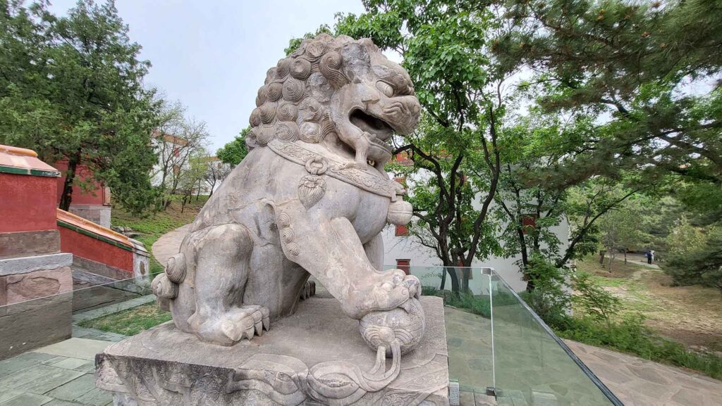 Seitenprofil eines großen, stolzen Steinlöwen mit gelockter Mähne, der seine Pfote auf einen verzierten Ball stützt.