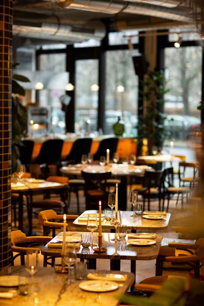 Stimmungsvolle Innenansicht des Restaurants Berta in Berlin mit Kerzenlicht auf gedeckten Marmortischen, warmem Licht, großen Fensterfronten und urban-gemütlicher Atmosphäre.