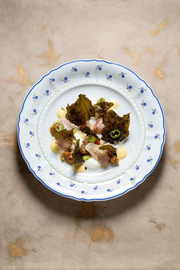 Feine Vorspeise im Berta Berlin auf floral verziertem Teller mit rohem Fisch, gefüllten Weinblättern, Olivenringen und cremigen Saucen, minimalistisch angerichtet.