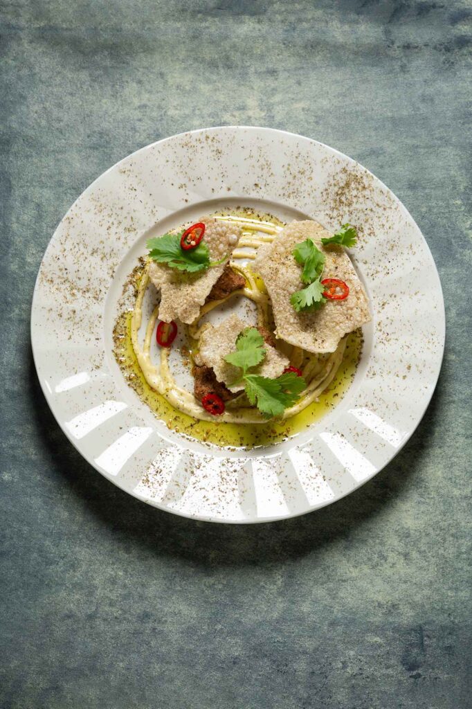 Kreativ angerichtetes Gericht im Berta Berlin mit knusprigen Elementen, frischen Kräutern, Chili und aromatischem Öl auf weißem Teller vor grauem Hintergrund.