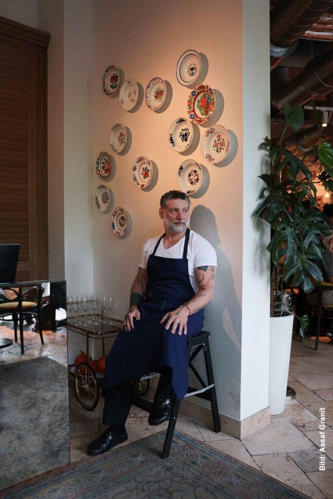 Start 1 Porträt von Assaf Granit im Restaurant Berta in Berlin, sitzend vor einer Wand mit dekorativen Tellern, in Kochschürze, mit ruhigem, konzentriertem Blick.