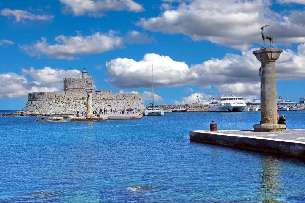 Mandraki-Hafen auf Rhodos mit der Festung Agios Nikolaos, Hirschstatuen auf Säulen, Booten im Wasser und blauem Himmel mit Wolken.