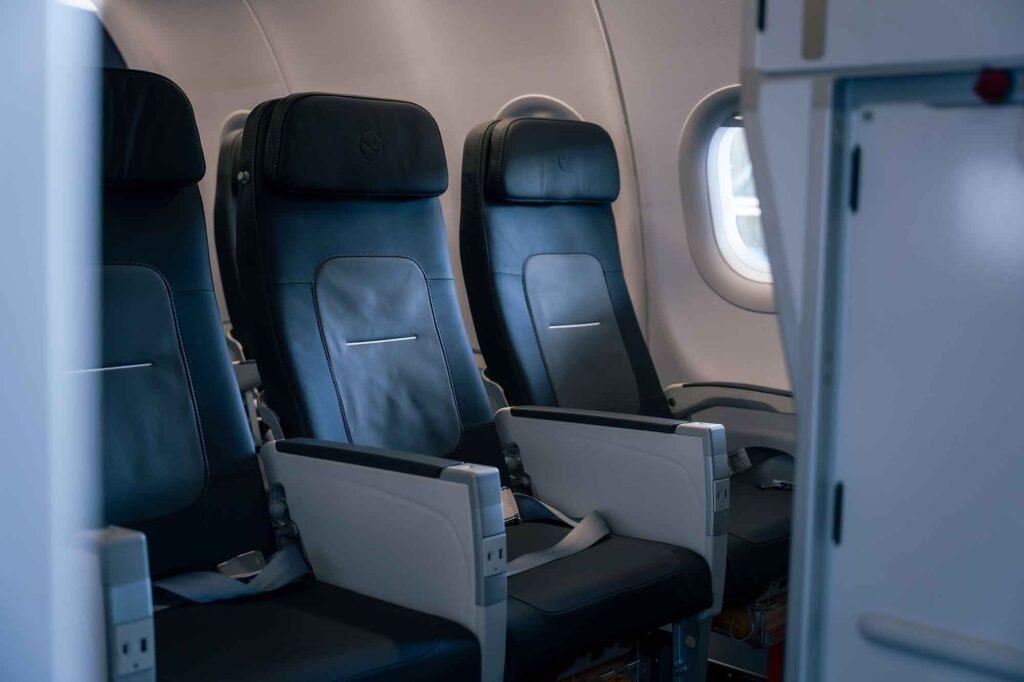 Moderne Economy-Class-Sitzreihe in einem Lufthansa Airbus A320 mit neuen dunklen Ledersitzen, verstellbaren Kopfstützen und heller Kabinenverkleidung.