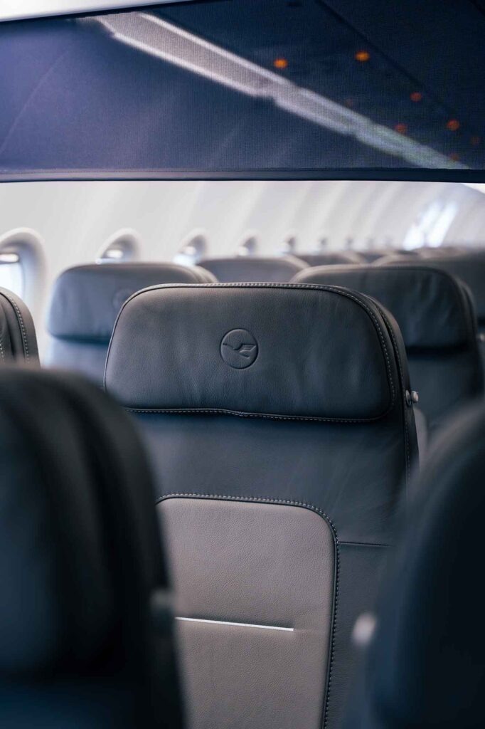 Detailaufnahme einer ergonomischen Kopfstütze aus dunkelblauem Leder mit dem Lufthansa-Kranich-Logo. Im unscharfen Hintergrund sind die weiteren Sitzreihen der modernisierten Kabine erkennbar.