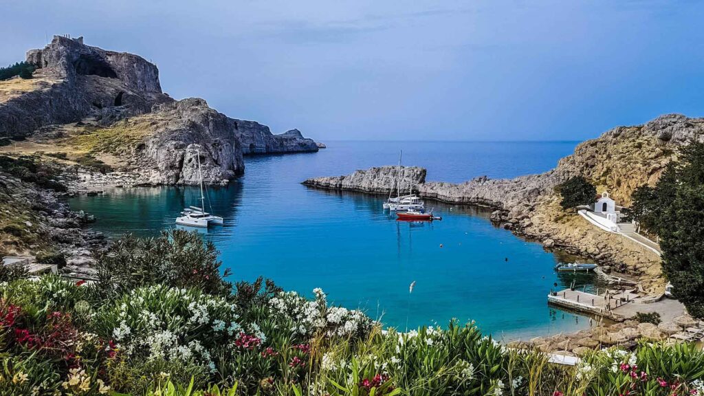 Türkisfarbene Bucht von Lindos auf Rhodos mit Segelbooten, felsigen Küsten, mediterraner Vegetation im Vordergrund und Blick zur Akropolis.