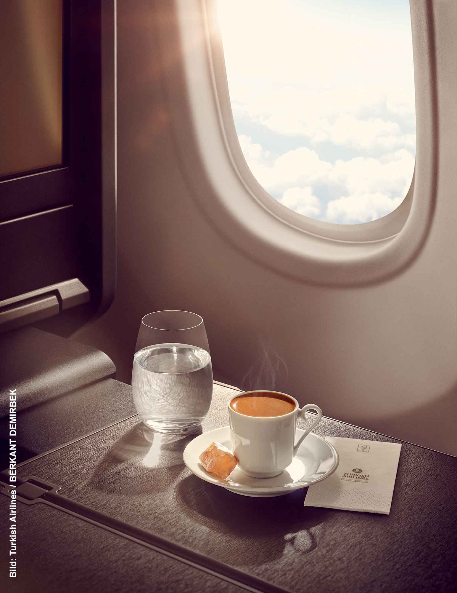 Eine Kaffee-Reise mit Turkish Airlines - Schluck für Schluck um die Welt 1 Tasse frisch servierter Kaffee auf einem Flugzeugtisch neben einem Glas Wasser und einem Stück Lokum, im Hintergrund das Flugzeugfenster mit Blick auf die Wolken.