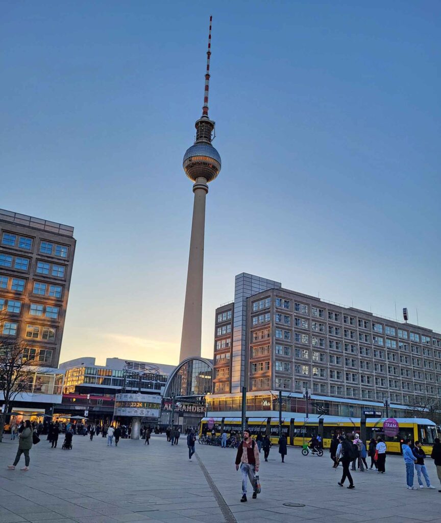 Berliner Fernsehturm am Alexanderplatz in der Dämmerung, umgeben von Gebäuden, Straßenbahn und Passanten.