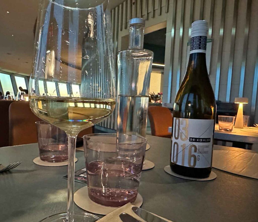 Gedeckter Tisch im Restaurant Sphere by Tim Raue mit Weißweinglas, Wasserkaraffe, Wasserflasche und Weinflasche in modernem Ambiente.