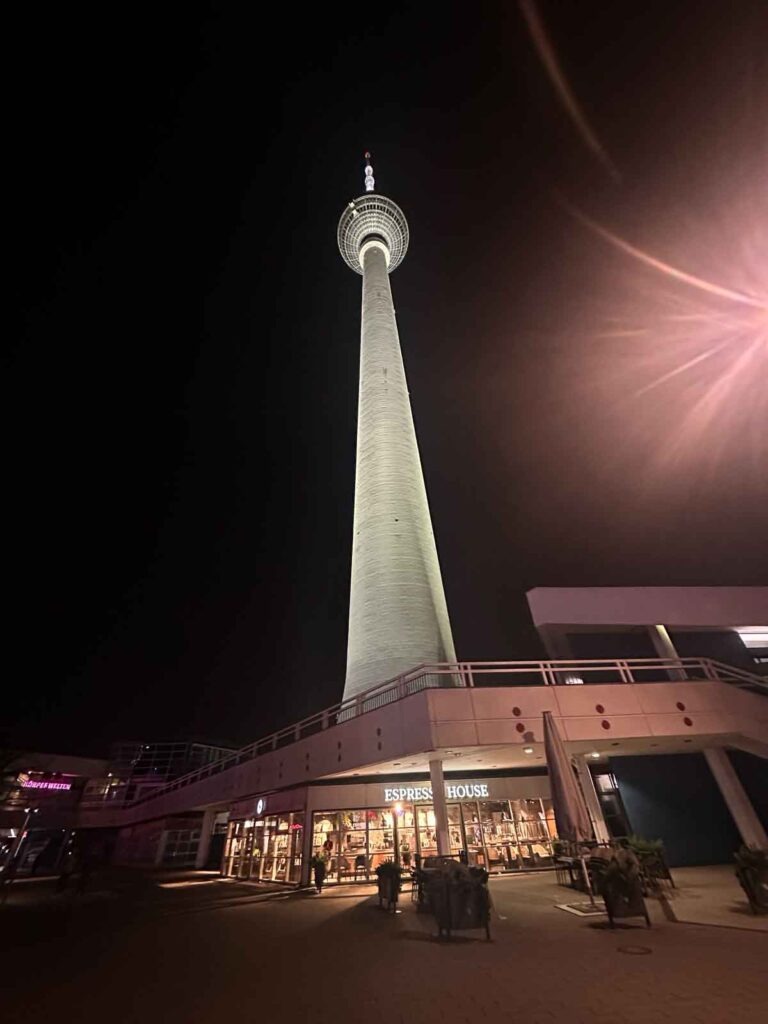 Nächtlicher Blick vom Fuß des Berliner Fernsehturms nach oben, darunter das beleuchtete Gebäude mit dem Espresso House am Alexanderplatz.