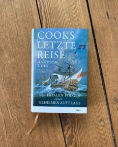 Buch-Tipp: Cooks letzte Reise – zwischen Genie, Gewalt und Grenzerfahrung 22 Buchcover von „Cooks letzte Reise“ von Hampton Sides mit Segelschiff auf stürmischer See auf Holzboden