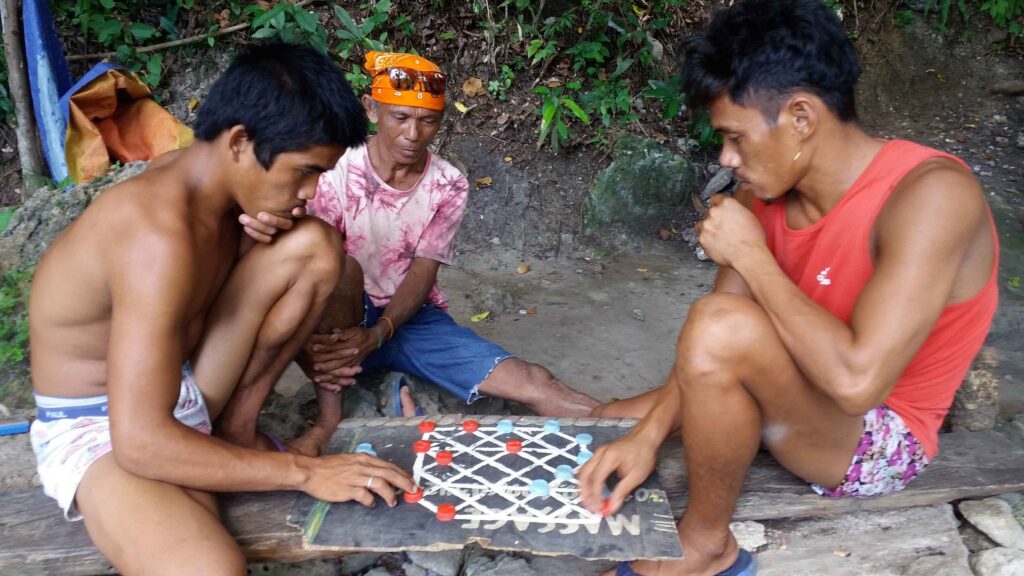 Drei Männer spielen ein einfaches Brettspiel mit roten und blauen Spielsteinen auf einem improvisierten Spielbrett.
