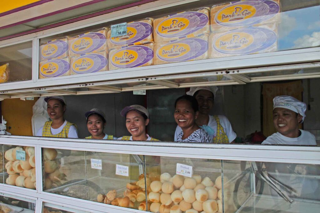 Mehrere Verkäuferinnen in einer kleinen Bäckerei mit frischen Brötchen hinter der Verkaufstheke.