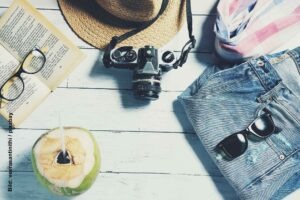 Sommerliches Reise-Stillleben mit Strohhut, analoger Kamera, Sonnenbrille, Jeans, Buch und frischer Kokosnuss auf weißem Holztisch.