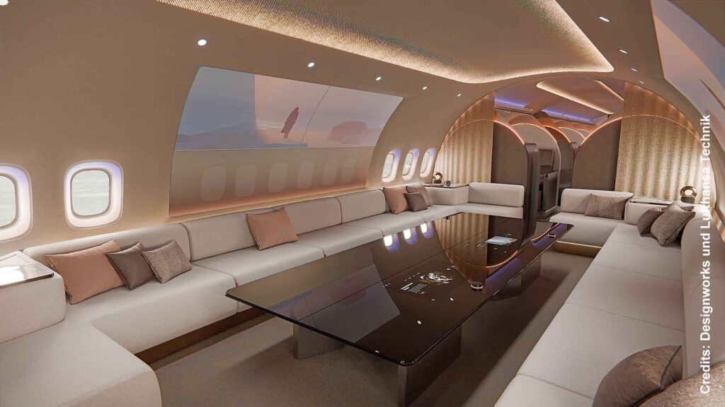 Eine großzügige Lounge-Sektion im Flugzeug mit umlaufenden, hellen Sofas und einem großen, dunklen Glastisch in der Mitte. An der Wand befindet sich ein riesiger Bildschirm.
