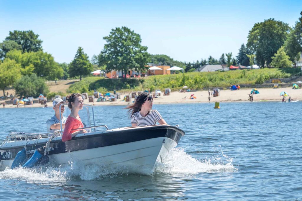 Lausitzer Seenland - Fünf-Seen-Verbund öffnet Ende Juni 6 Drei Personen fahren mit einem Motorboot über den Senftenberger See, im Hintergrund ein belebter Sandstrand mit Badegästen