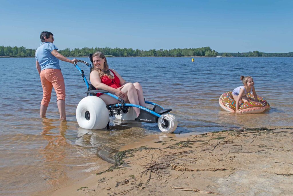Lausitzer Seenland nach Reisen für Alle zertifiziert 3 Frau sitzt in einem Amphibienrollstuhl im flachen Wasser des Senftenberger Sees, begleitet von einer Person, während ein Kind auf einem Schwimmring spielt