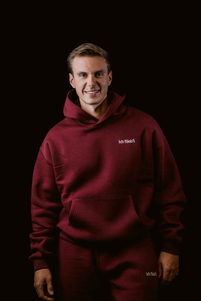 Eurowings: Was sonst noch wichtig ist 19 Mann trägt roten Kapuzenpullover und Jogginghose mit „Ich flieb’s“-Logo vor dunklem Hintergrund