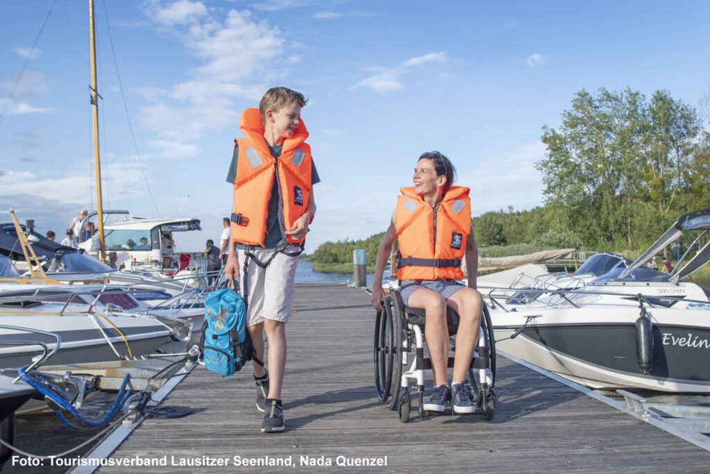 Junge und Frau im Rollstuhl mit Schwimmwesten gehen über einen Steg zu einer Marina mit Booten am Senftenberger See