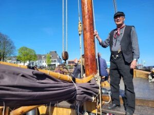 Ein älterer Mann mit Brille, dunkler Mütze, gestreiftem Hemd, schwarzer Weste und rotem Halstuch steht lächelnd an Deck eines Holzschiffes neben dem Holzmast vor blauem Himmel.