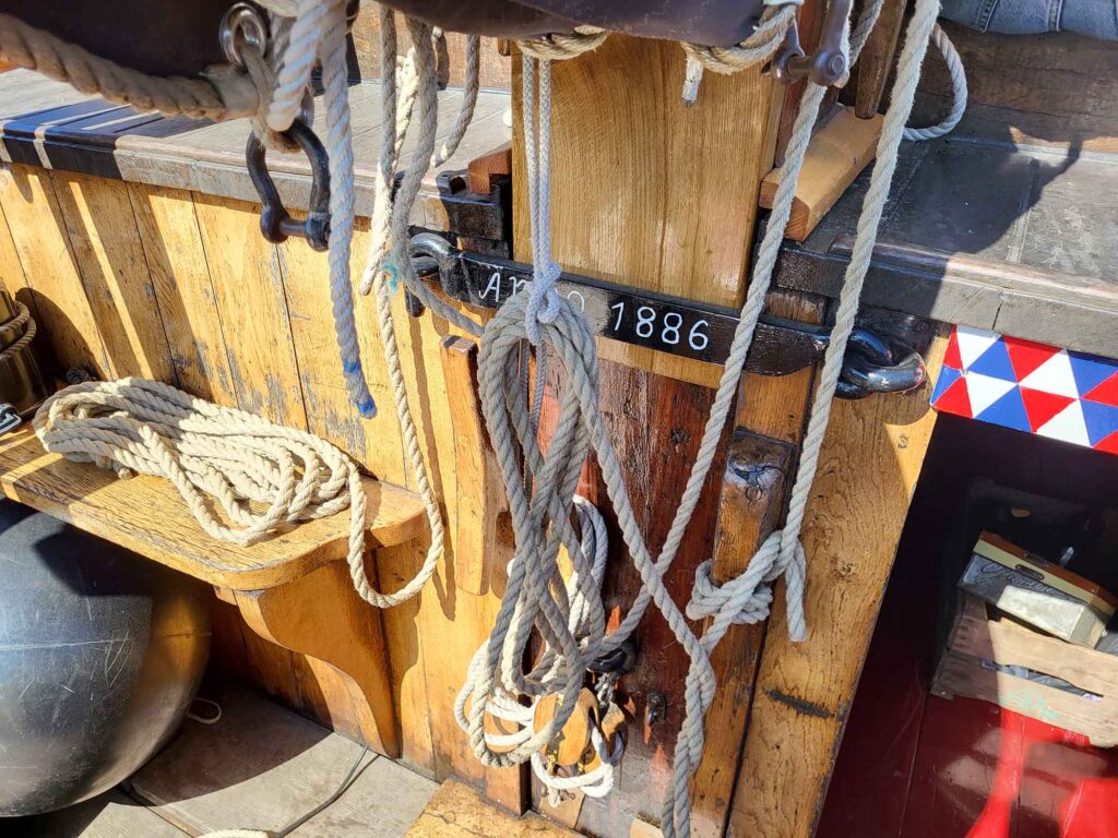 Maritimes Erbe erleben: Eine traditionelle Botterfahrt auf dem Veluwemeer ab Harderwijk 3 Nahaufnahme von Seilen und Taubeschlägen an einem hölzernen Schiffsmast. Auf einer schwarzen Metallschiene steht in weißer Schrift „Anno 1886“.