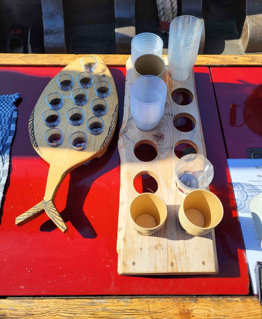 Maritimes Erbe erleben: Eine traditionelle Botterfahrt auf dem Veluwemeer ab Harderwijk 9 Draufsicht auf einen roten Tisch an Deck, auf dem ein fischförmiges Holzbrett mit gefüllten Schnapsgläsern und Halterungen für Becher steht.