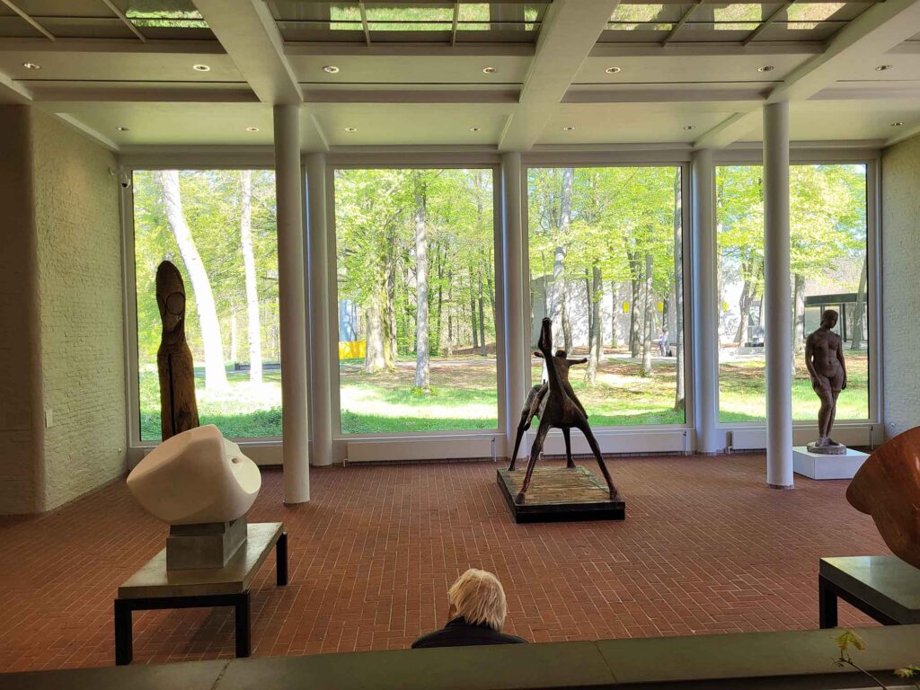 Heller Museumsraum mit großen Fensterfronten und modernen Skulpturen, dahinter ein parkähnliches Gelände mit Bäumen