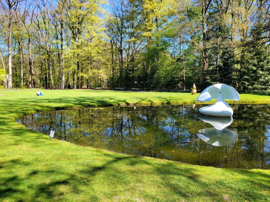 Moderne weiße Skulptur in einem Teich mit Spiegelung, umgeben von Wiese und Bäumen im Skulpturenpark