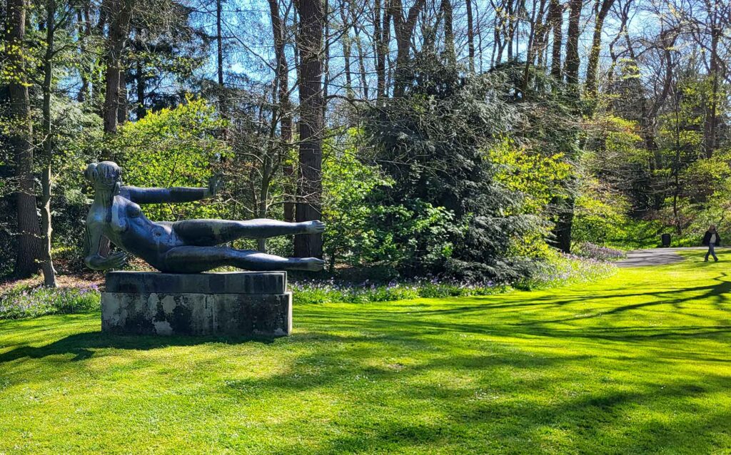 Bronzeskulptur einer liegenden weiblichen Figur auf einem Sockel im grünen Parkgelände