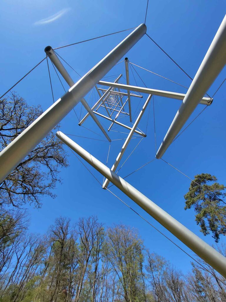 Blick von unten durch eine geometrische Stahlkonstruktion in den blauen Himmel mit umliegenden Bäumen