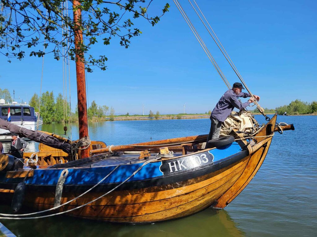 Maritimes Erbe erleben: Eine traditionelle Botterfahrt auf dem Veluwemeer ab Harderwijk 4 Seitenansicht des hölzernen Botters mit der Aufschrift „HK 103“ am Bug, während ein Seemann auf dem Vorschiff an den Seilen arbeitet, im Hintergrund blaues Wasser und Windräder.