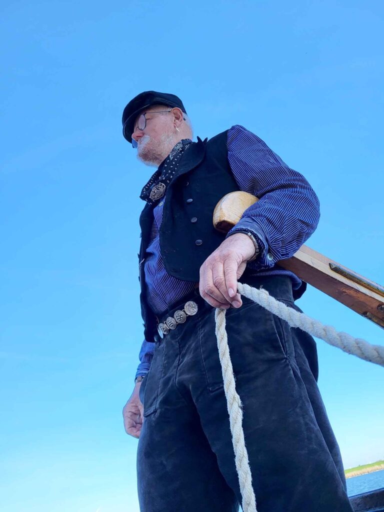 Maritimes Erbe erleben: Eine traditionelle Botterfahrt auf dem Veluwemeer ab Harderwijk 6 Froschperspektive eines älteren Seemanns mit grauem Bart, Brille und Schiffermütze, der eine Leine hält und den Blick vor strahlend blauem Himmel in die Ferne richtet.