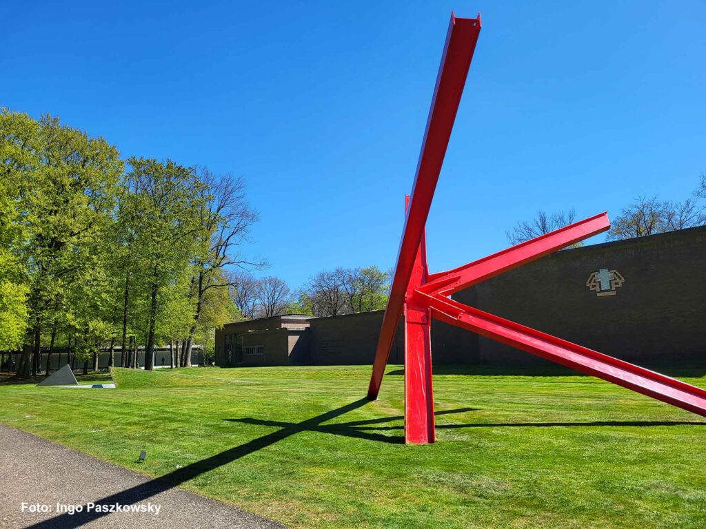 Große abstrakte rote Stahlskulptur auf einer Wiese vor dem modernen Museumsbau
