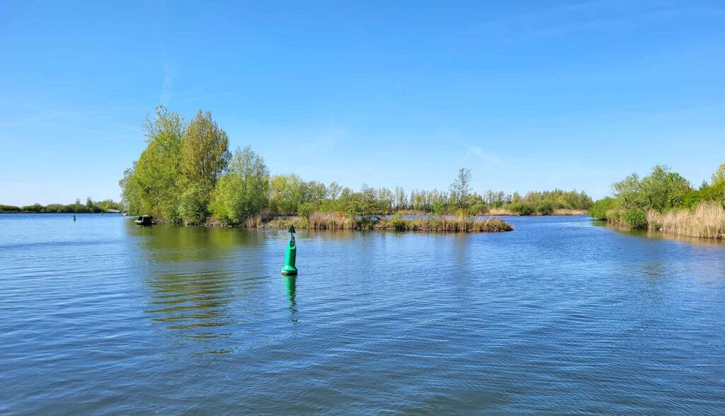 Maritimes Erbe erleben: Eine traditionelle Botterfahrt auf dem Veluwemeer ab Harderwijk 8 Blick über ruhiges, blaues Wasser auf eine kleine, dicht bewachsene grüne Insel unter wolkenlosem blauem Himmel, mit einer grünen Fahrwassertonne im Vordergrund.