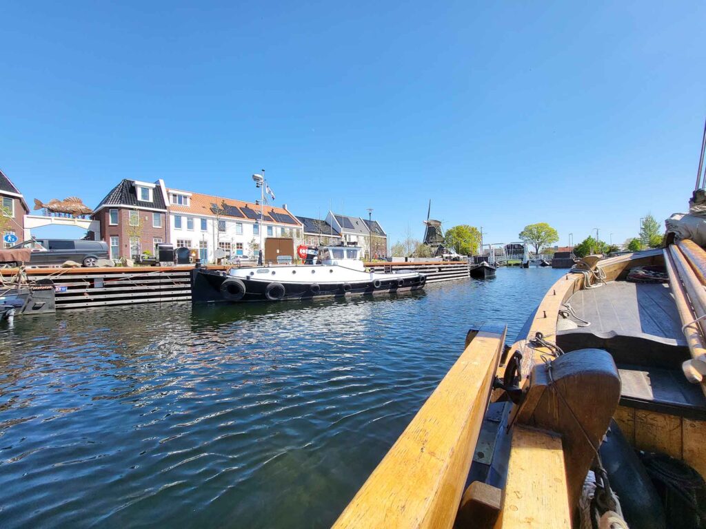 Maritimes Erbe erleben: Eine traditionelle Botterfahrt auf dem Veluwemeer ab Harderwijk 7 Perspektive vom Holzdeck des Botters entlang eines Kanals in Harderwijk. Auf der linken Seite liegen ein schwarz-weißes Arbeitsboot und moderne Wohnhäuser am Kai. Im Hintergrund sind eine historische Windmühle und eine Zugbrücke unter strahlend blauem Himmel zu erkennen.