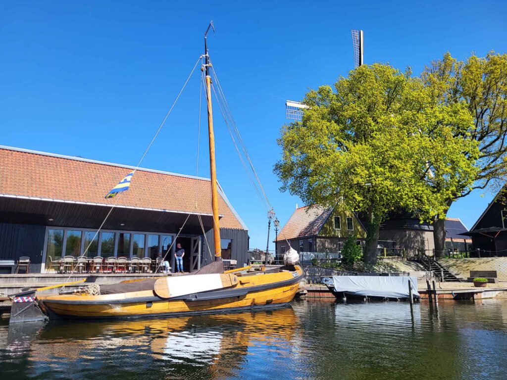 Maritimes Erbe erleben: Eine traditionelle Botterfahrt auf dem Veluwemeer ab Harderwijk 5 Ein hölzernes Segelschiff liegt vertäut an einem Steg vor einem modernen, schwarzen Pavillon mit Glasfront. Rechts daneben rahmen ein großer, grüner Baum und eine traditionelle holländische Windmühle die Szenerie ein. Das Schiff wirft eine klare Spiegelung auf die Wasseroberfläche.