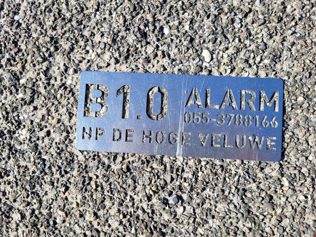 Metallplakette mit Aufschrift „NP De Hoge Veluwe“ auf einem Asphaltweg im Nationalpark
