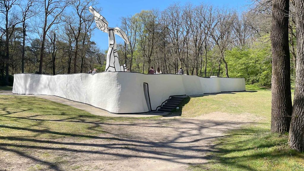 Moderne Skulptur auf erhöhter Plattform im Skulpturenpark des Kröller-Müller Museums, umgeben von Bäumen und Spazierwegen