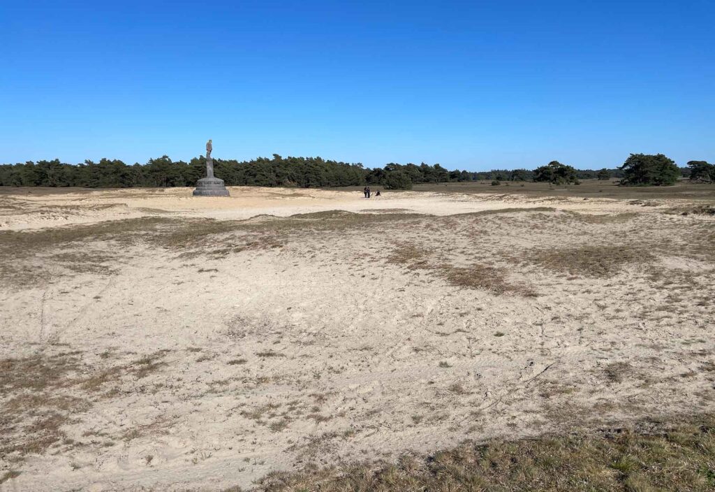 Weite, sandige Heidelandschaft im Nationalpark De Hoge Veluwe mit vereinzelten Bäumen und Denkmal in der Ferne