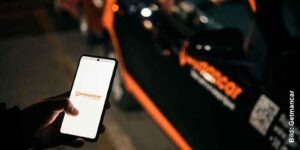 Hand hält Smartphone mit Carsharing-App vor geparktem Auto im Dunkeln mit orange beleuchtetem Logo