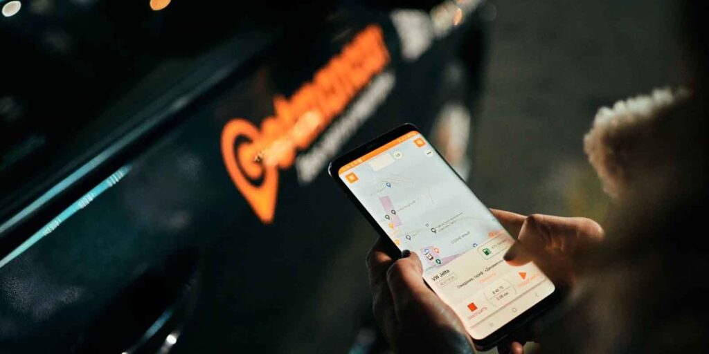 Nutzer bedient Smartphone mit Kartenansicht einer Carsharing-App neben Fahrzeug mit Logo im Hintergrund