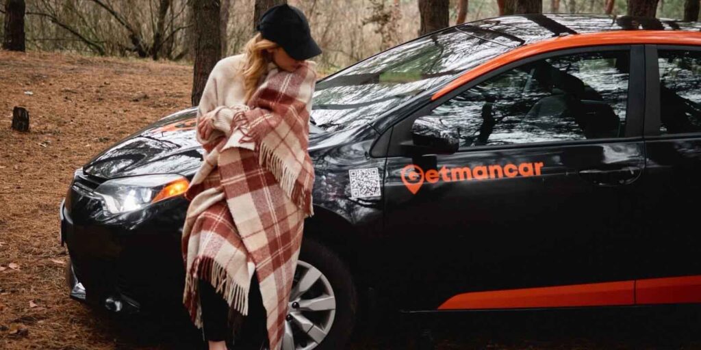 Person mit Decke steht neben schwarzem Carsharing-Auto im Wald bei kühlem Wetter