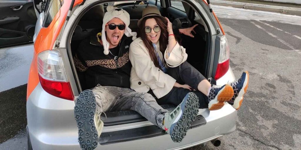 Zwei junge Erwachsene sitzen lachend im offenen Kofferraum eines Autos und genießen einen Ausflug
