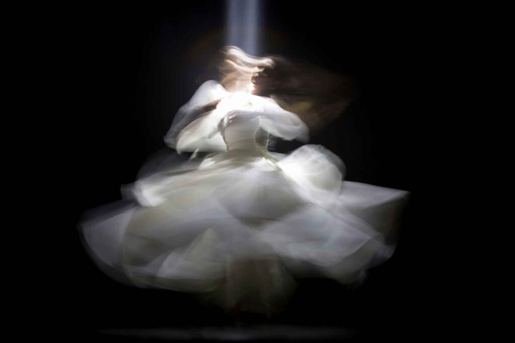 Wie sind Ballettkunst und digitale Technologien miteinander verbunden? 3 Ballerina in weißem Kleid dreht sich im Scheinwerferlicht auf dunkler Bühne, ihr Gesicht ist durch Bewegungsunschärfe kaum erkennbar.