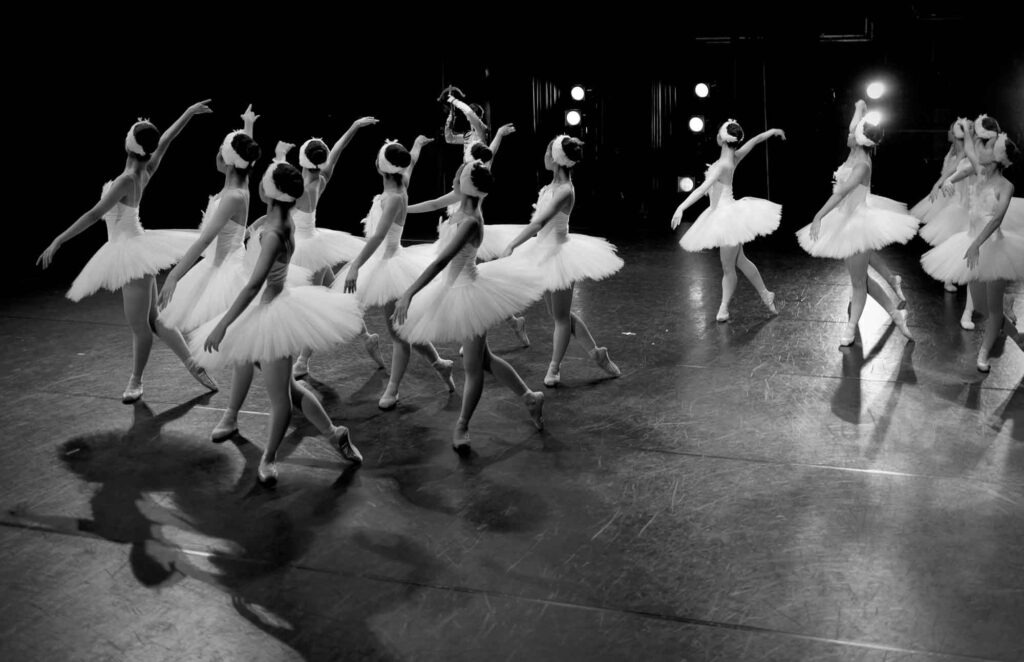 Wie sind Ballettkunst und digitale Technologien miteinander verbunden? 4 Ballettgruppe in weißen Tutus tanzt auf einer dunklen Bühne in Schwarz-Weiß.