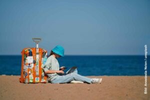 Sicher unterwegs arbeiten als digitaler Nomade 13 Reisende mit Laptop und Smartphone sitzt neben einem orangefarbenen Koffer am Strand und arbeitet vor dem Meer.