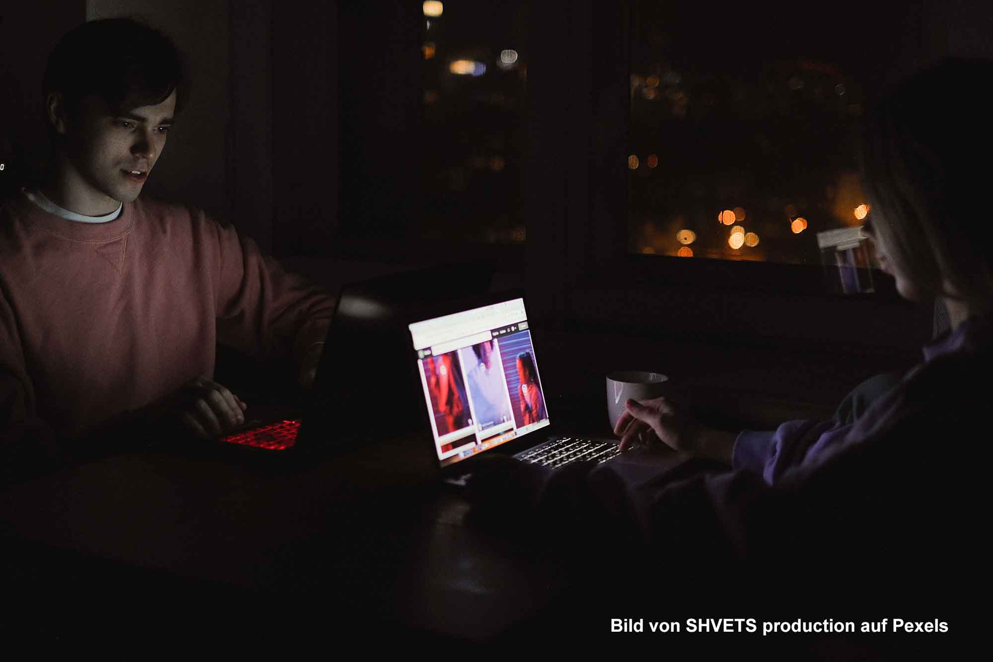 Zwei Personen sitzen nachts mit geöffneten Laptops in einem dunklen Raum.