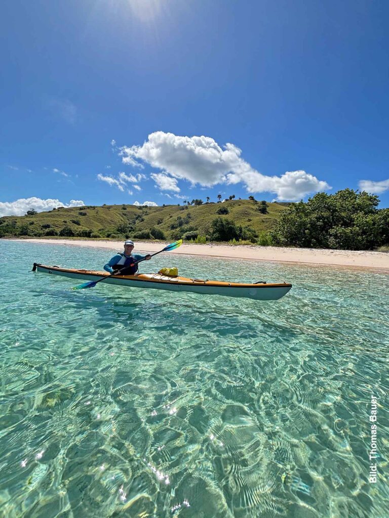 Thomas Bauer in einem Seekajak auf glasklarem Wasser vor einem hellen Sandstrand und grünen Hügeln